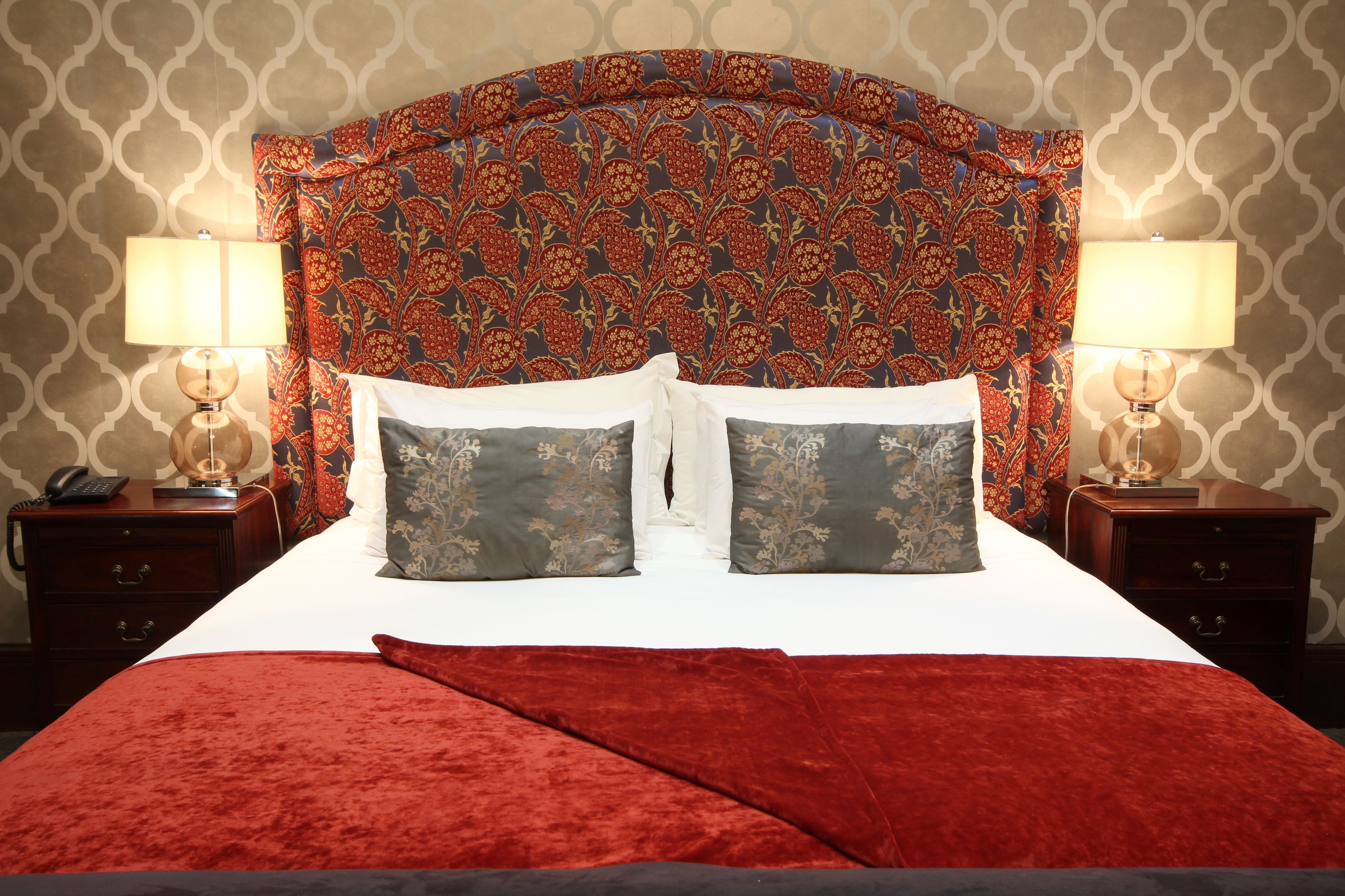 Un lit avec une couverture rouge, deux oreillers et une tête de lit à motifs. Il y a deux tables de chevet, chacune avec une lampe, et un téléphone sur l’une d’elles.
