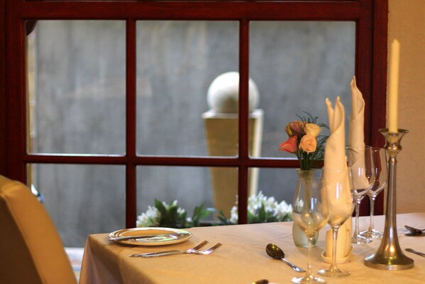 Una mesa de comedor dispuesta con platos, cubiertos y copas de vino, con una vela y flores al fondo.