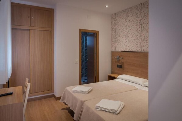A bedroom with two beds, a wooden wardrobe, a small table, and a door leading to a staircase. - San Antonio (Madrid)