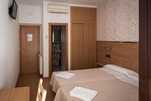 A hotel room with a bed, a wooden wardrobe, a radiator, a television on the wall, and a door with a sign. - San Antonio (Madrid)