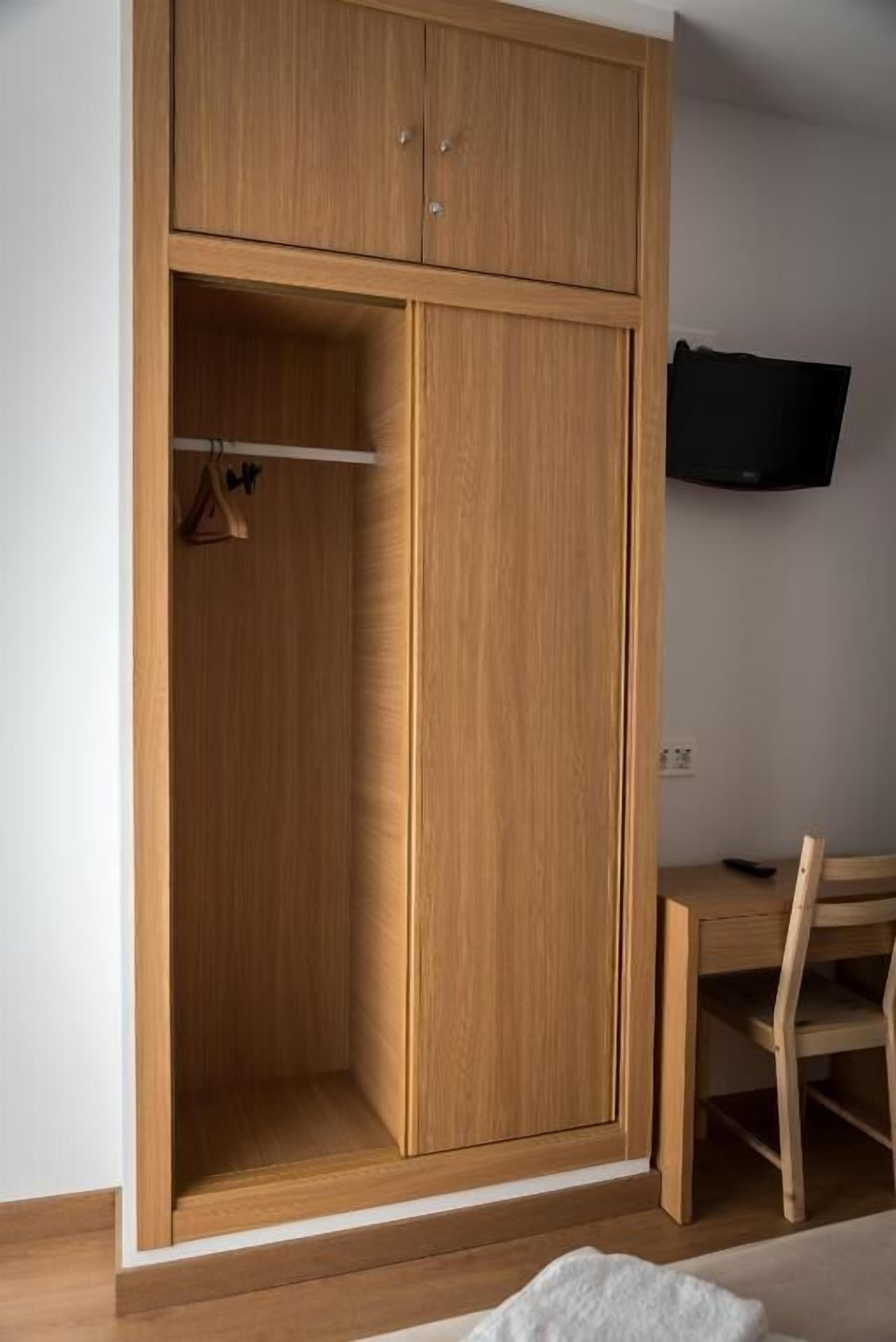 A wooden wardrobe with a hanging rod, a desk, and a chair in a room with a flat-screen TV mounted on the wall.