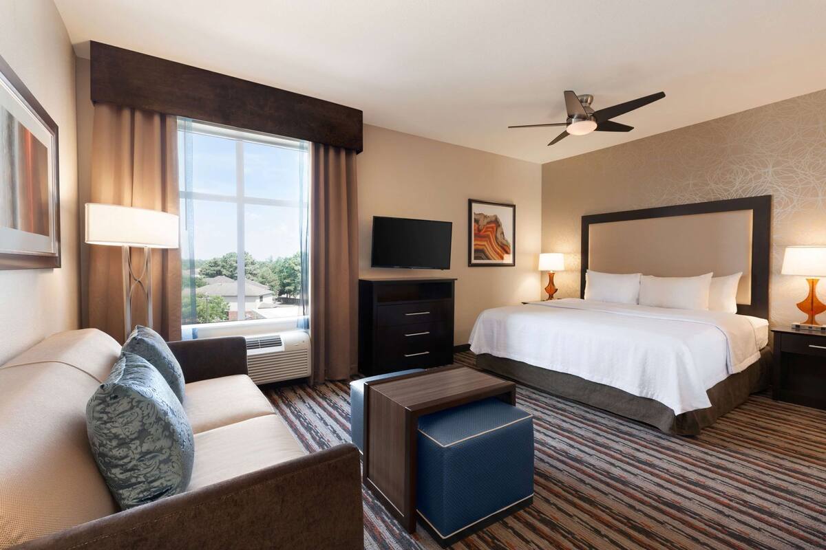 Habitación de hotel con cama, escritorio, silla, televisor y ventana con cortinas.