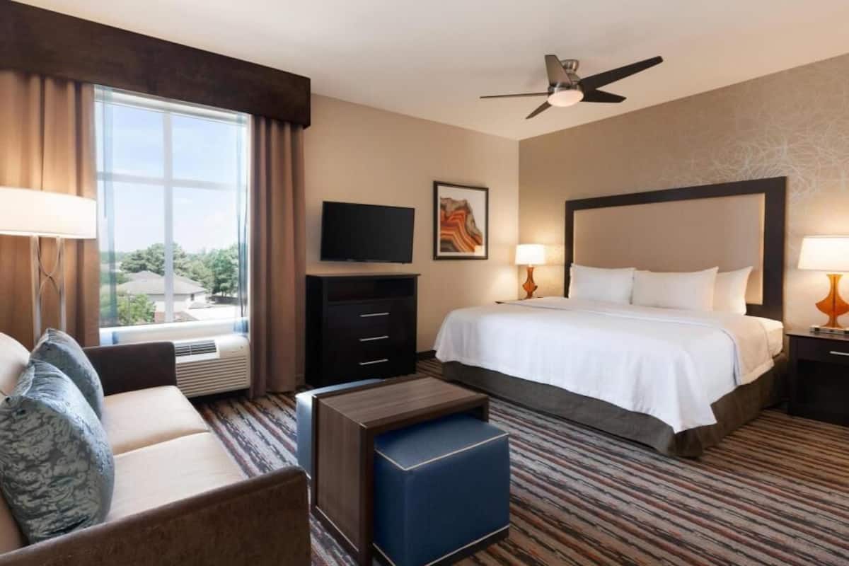 A hotel room with a bed, a sofa, a TV, a ceiling fan, and a window with curtains.