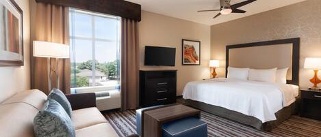 Habitación de hotel con cama, sofá, televisor, ventilador de techo y ventana con cortinas.