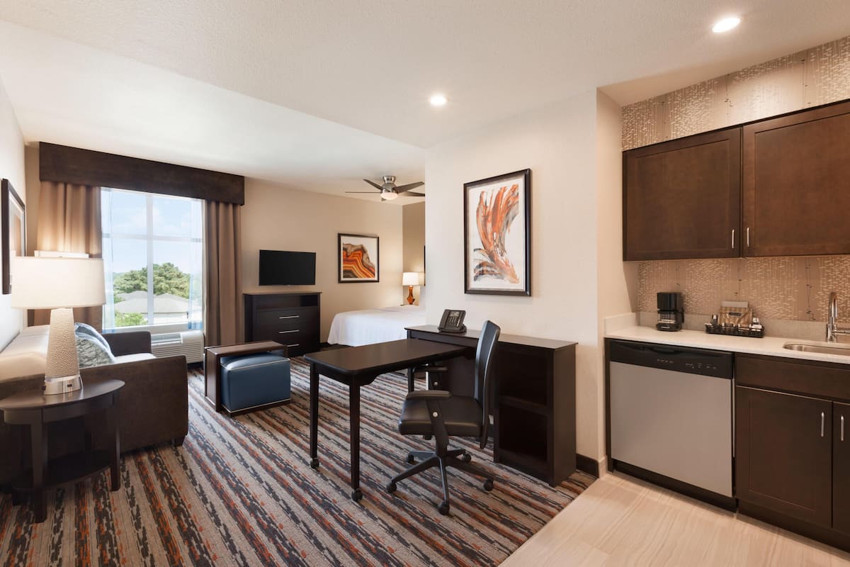 A hotel room with a bed, desk, chair, kitchenette, and large windows.