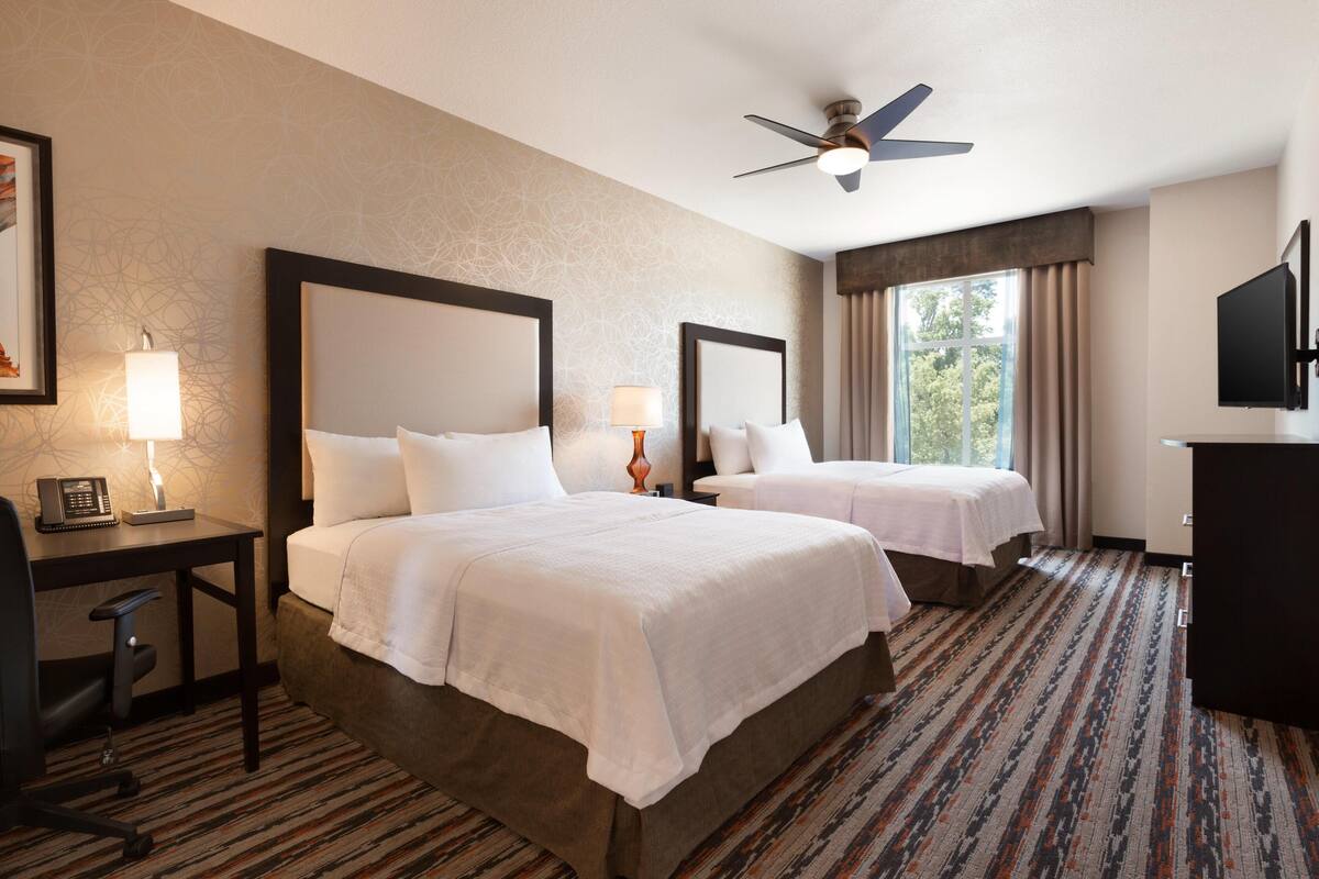 A hotel room with two beds, a desk with a telephone, a desk lamp, a ceiling fan, a TV, and a window with curtains.