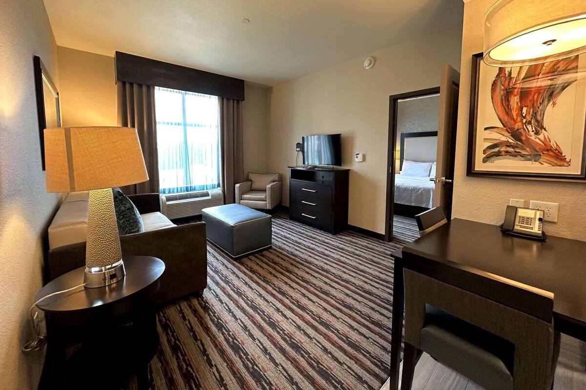 Habitación de hotel con sofá, escritorio, silla, televisor, cómoda, lámpara y un cuadro en la pared.