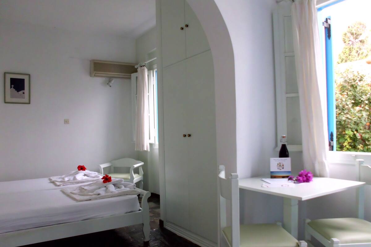 A small, clean room with a single bed, a white desk, and a chair. There is a window with curtains, a bottle on the desk, and a card on the wall.