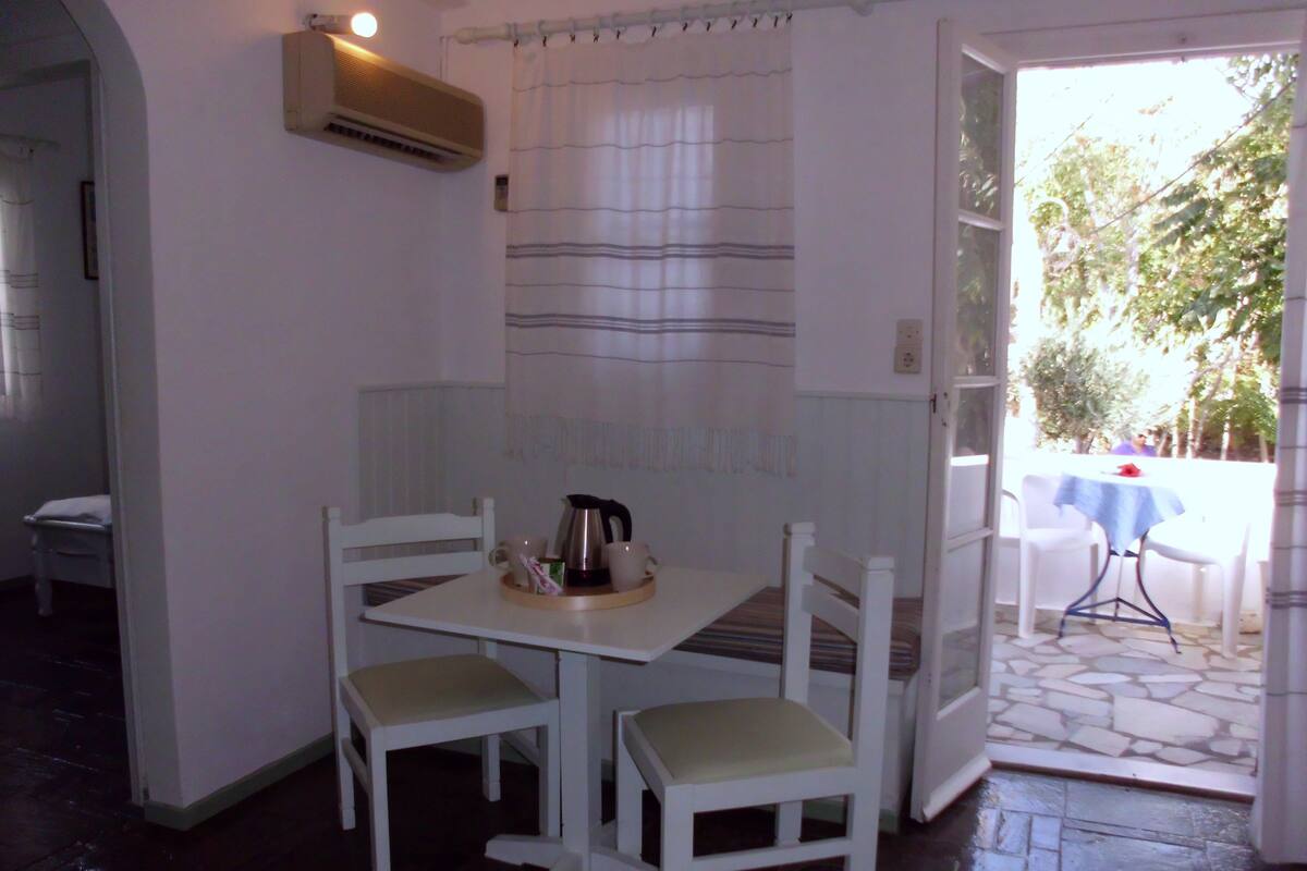 A small, cozy room with a table and two chairs, a kettle, and a window with curtains.