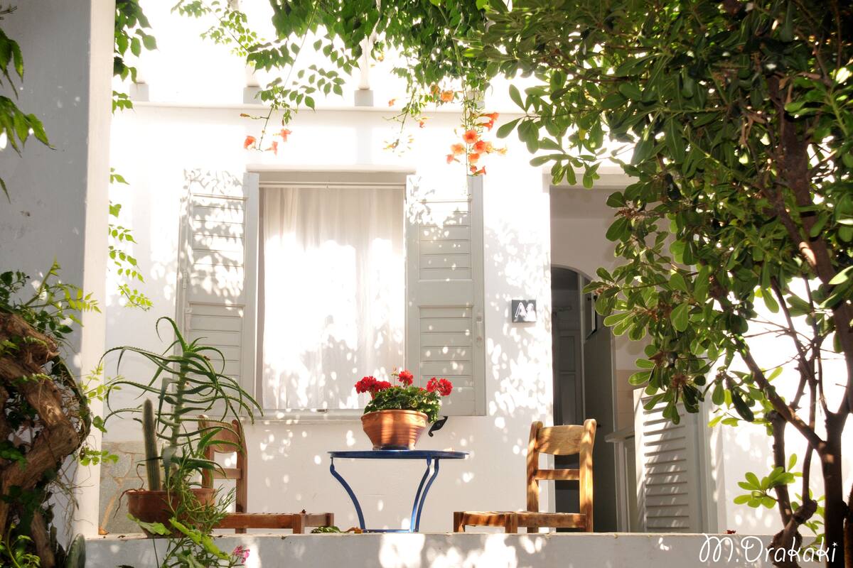 A white building with a window and shutters, a table with a copper pot of flowers, and two wooden chairs.