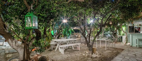 An outdoor dining area with tables and benches, surrounded by trees and illuminated by hanging lights.