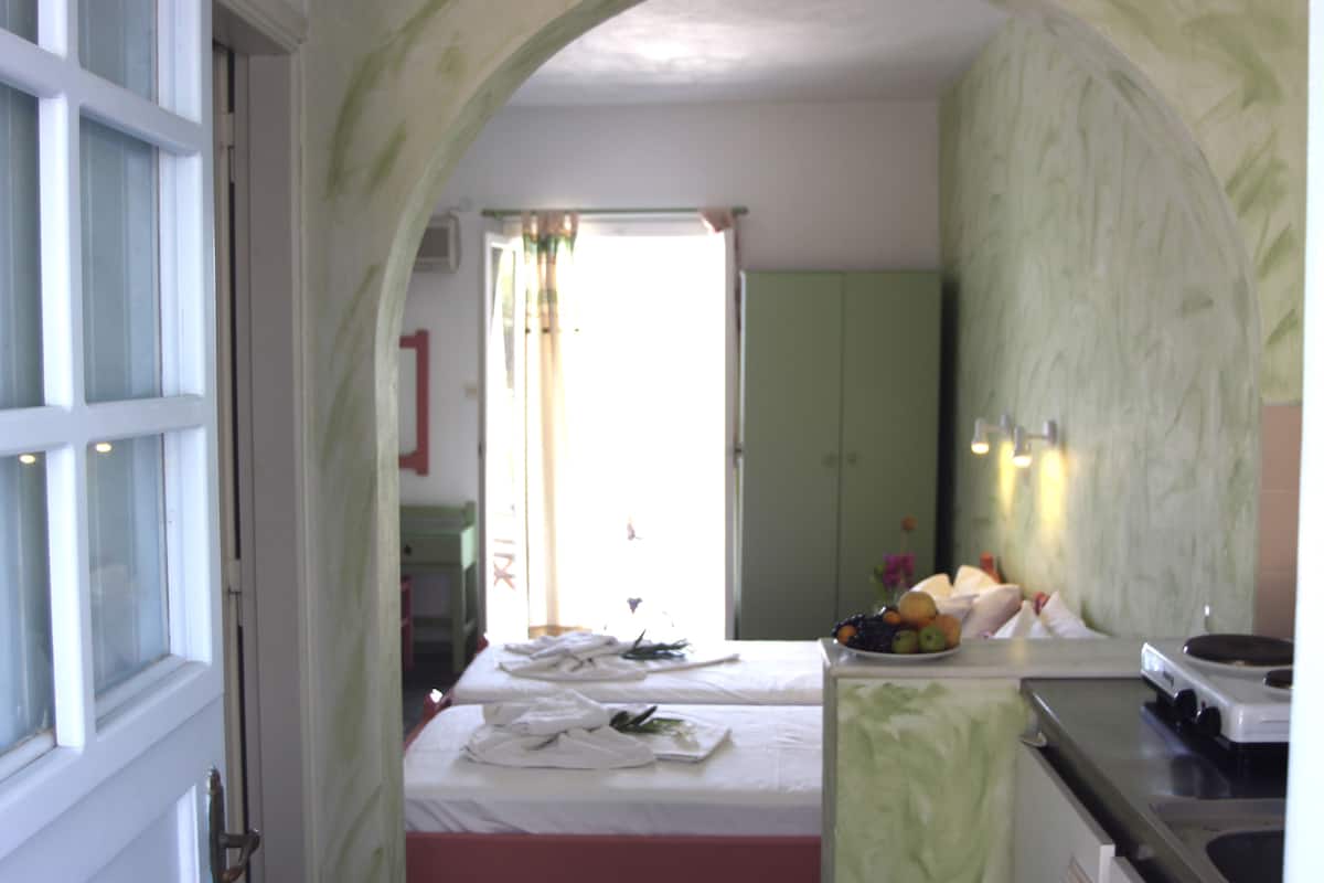 Un dormitorio con cama, una mesita de noche con frutero, ventana con cortinas y un armario verde.
