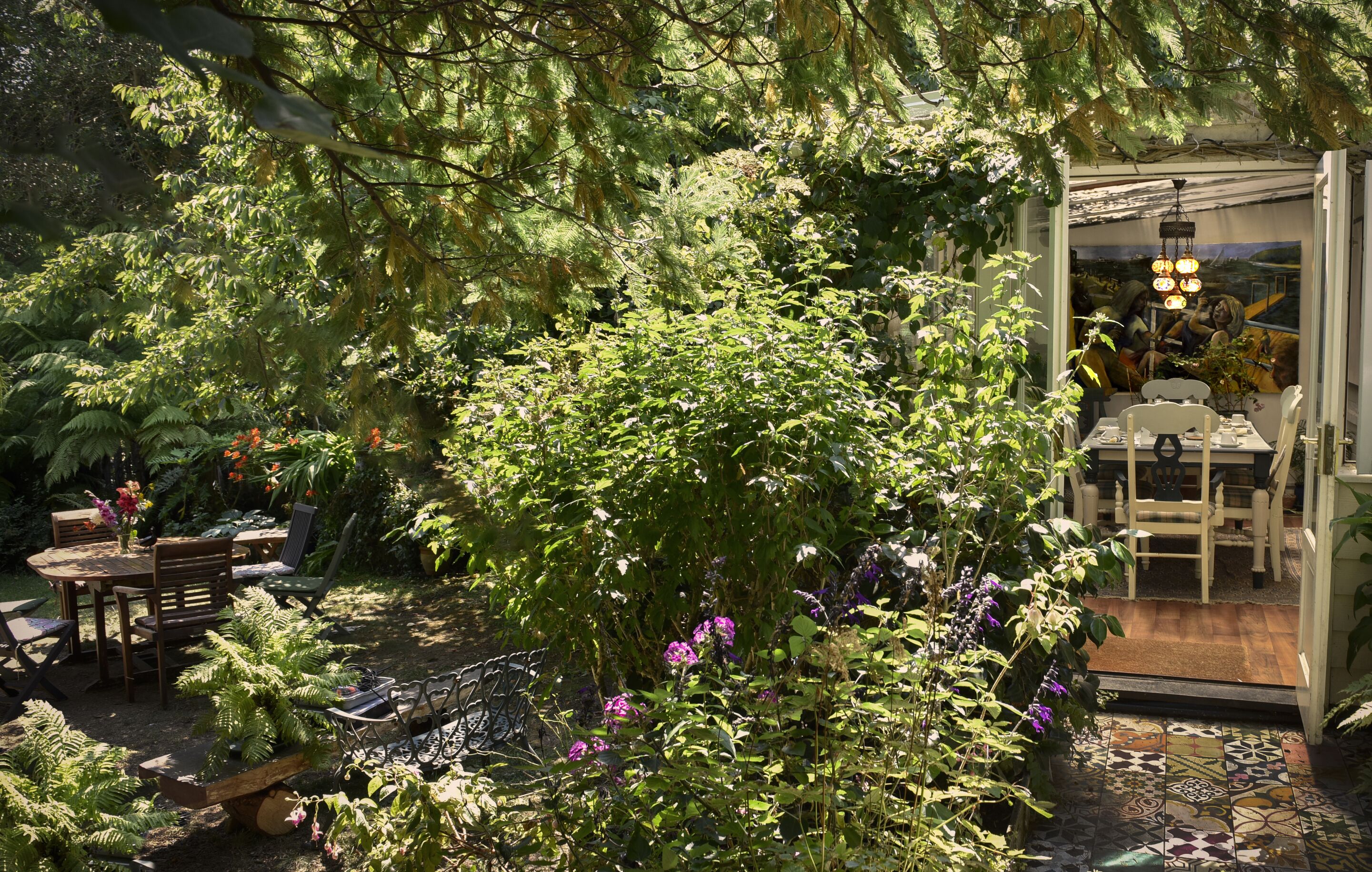 Een tuin met een eethoek, met tafels en stoelen, omringd door weelderige begroeiing en bloeiende planten.