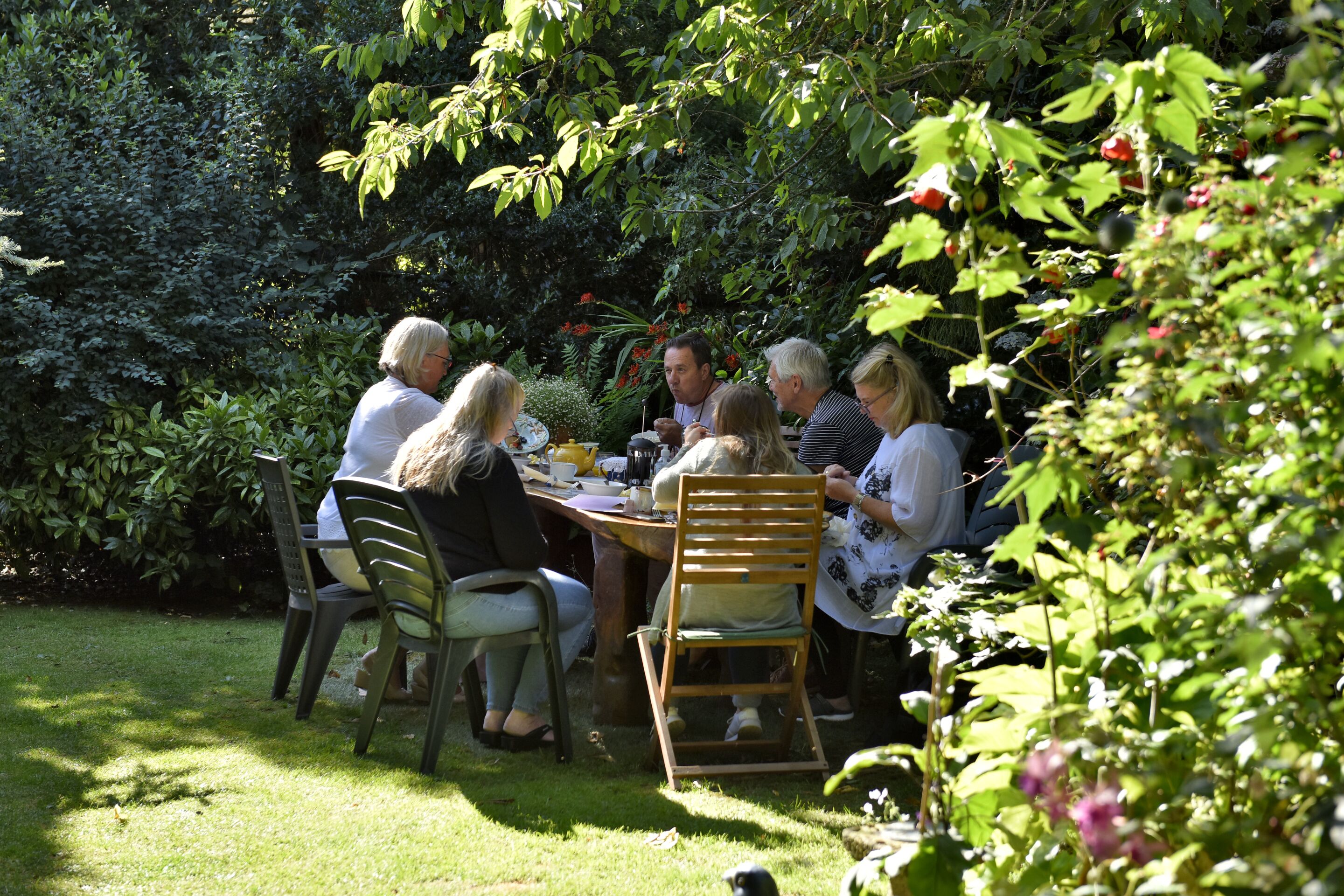 Un grupo de personas sentadas alrededor de una mesa en un jardín, con follaje y plantas al fondo.