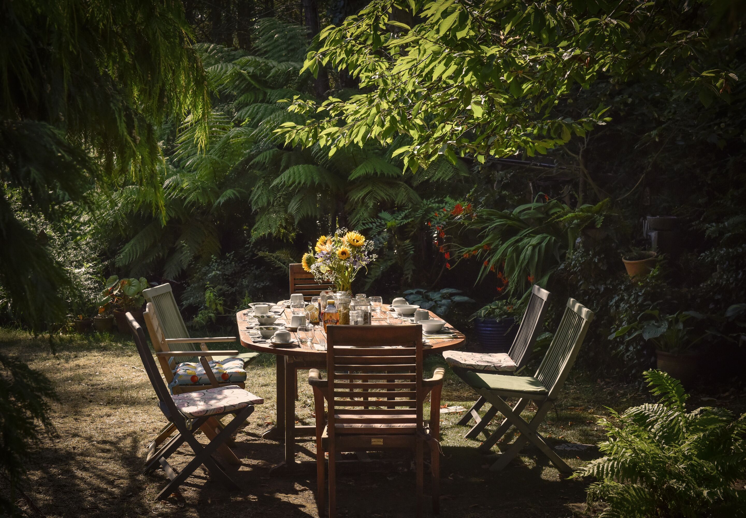 Un área de comedor al aire libre con una mesa redonda dispuesta para cuatro personas, rodeada de sillas, en un exuberante jardín.