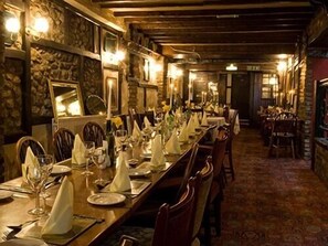 A long dining table set with white tablecloths, plates, and glasses, surrounded by wooden chairs in a warmly lit room with stone walls and a patterned carpet. - Dun Cow Inn (Stockton-on-Tees)