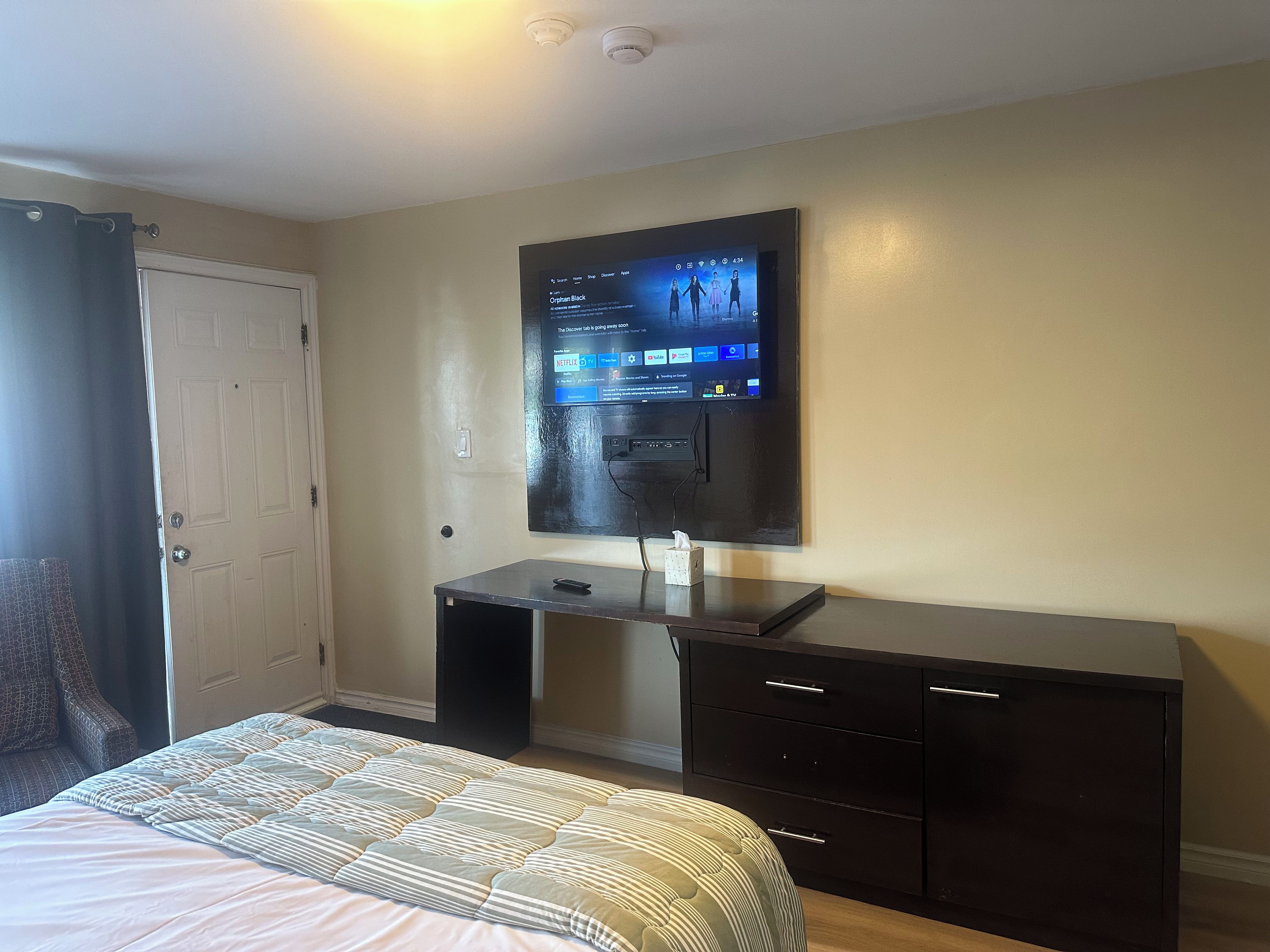 A hotel room with a bed, a desk with a computer, a television mounted on the wall, and a door.