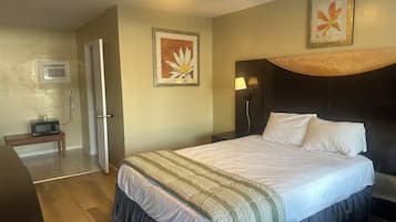 Ein Hotelzimmer mit einem Bett, einem Nachttisch, einer Lampe, einem Bilderrahmen und einer Mikrowelle auf einem kleinen Tisch.