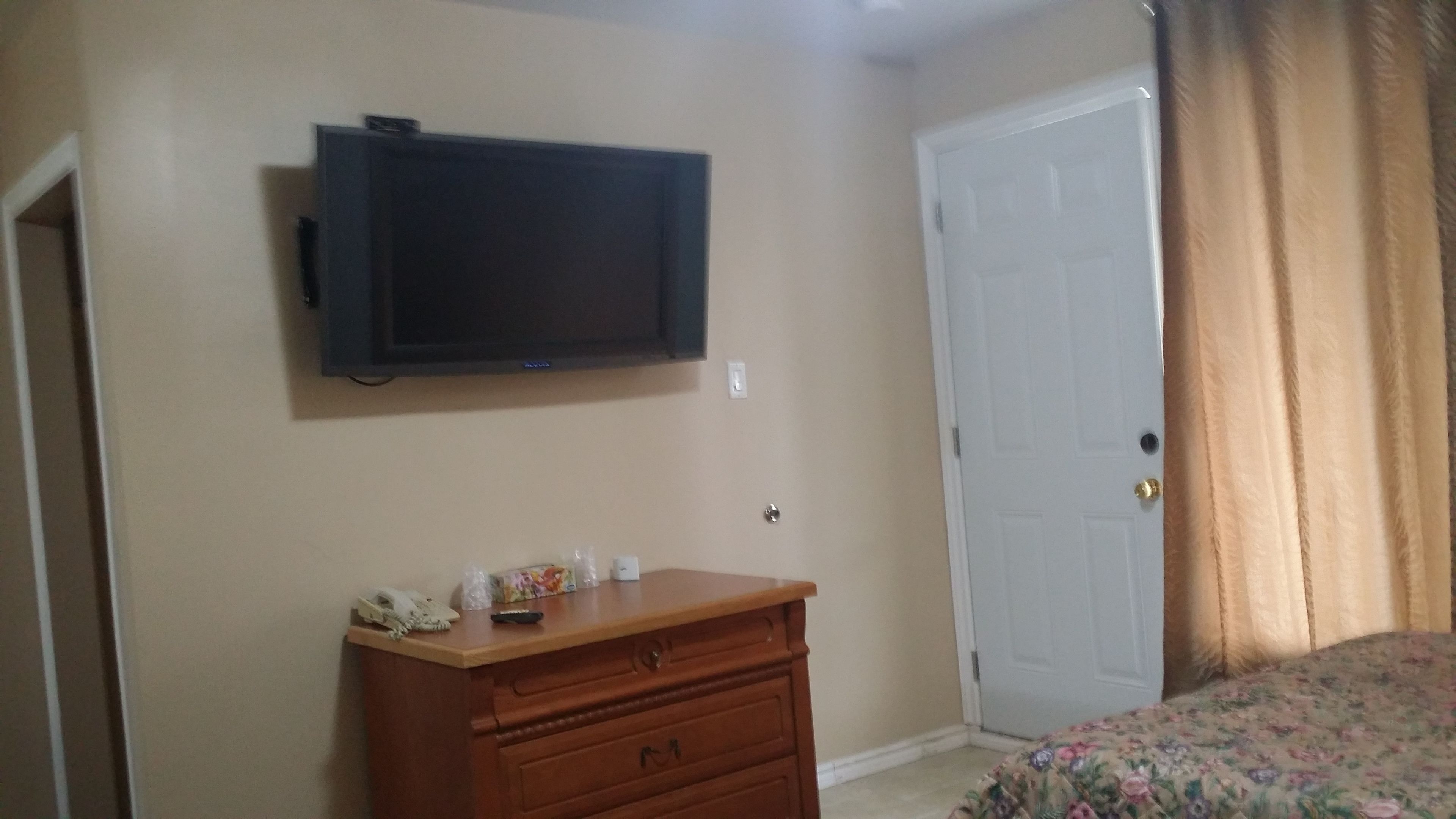 A bedroom with a television mounted on the wall, a wooden dresser, a door, and a bed with a floral bedspread.
