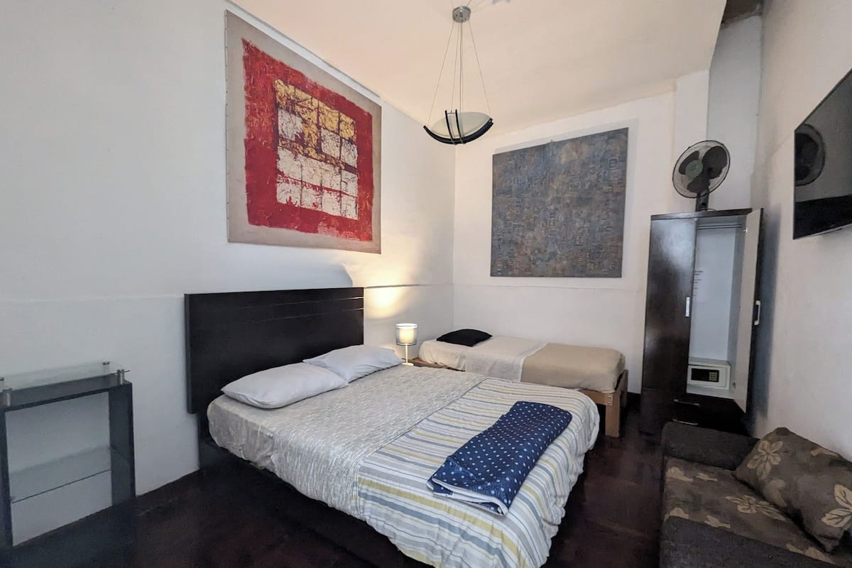 A bedroom with a bed, bedside tables, a nightstand, a wall-mounted fan, and artwork.