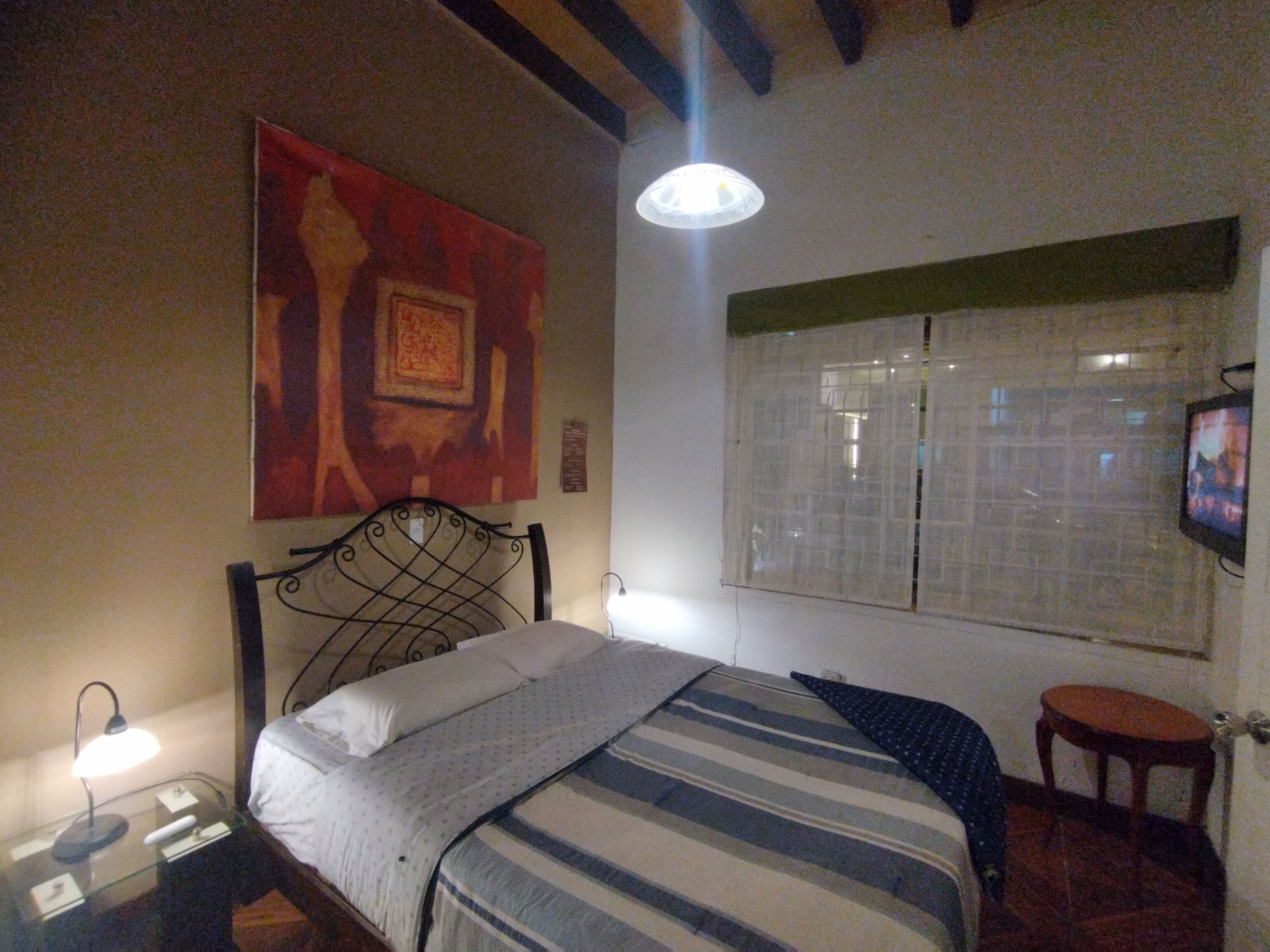A bedroom with a bed, a nightstand, a lamp, a painting on the wall, a window with blinds, and a television on the wall.