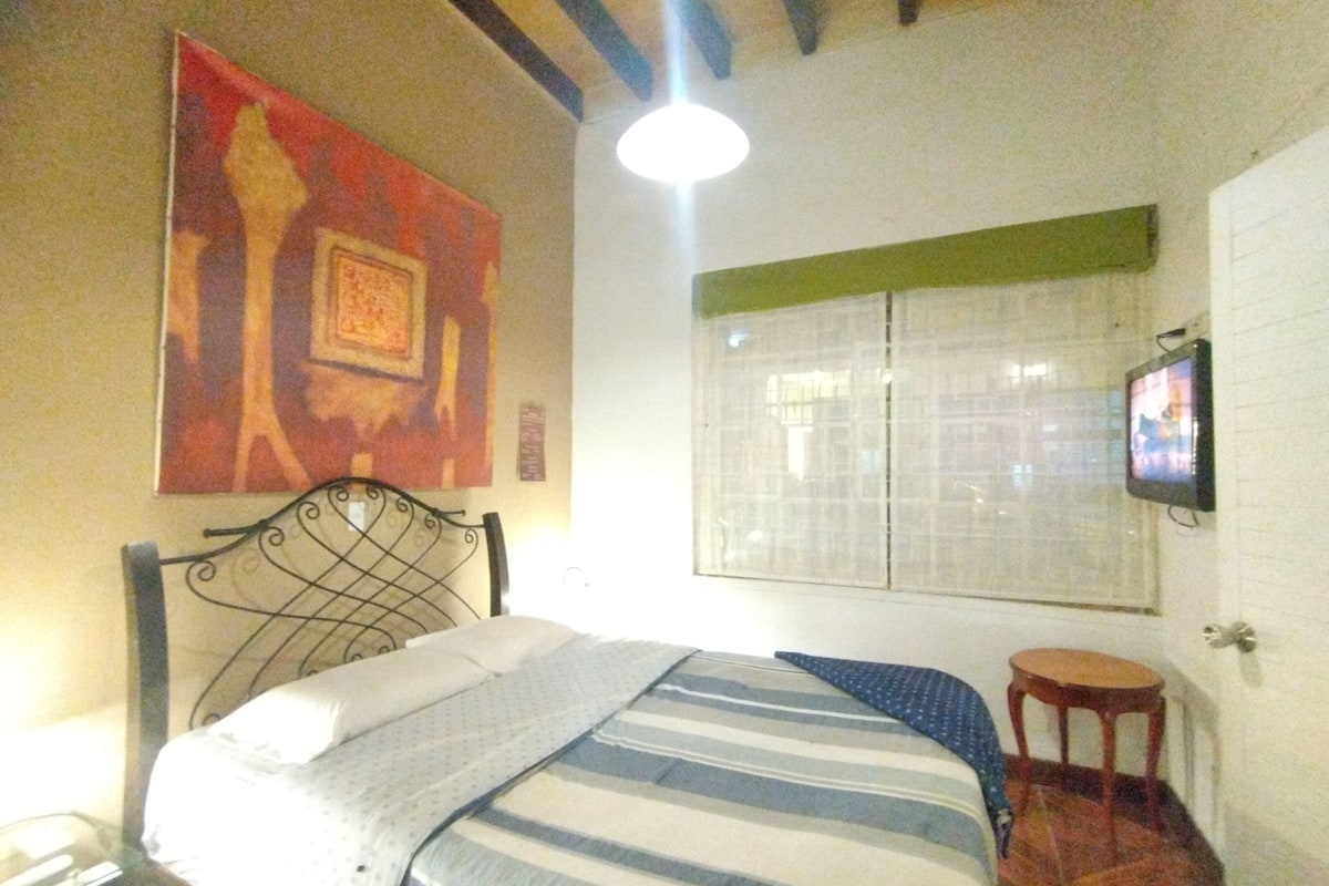 A bedroom with a bed, a television mounted on the wall, a window with blinds, and a painting on the wall.