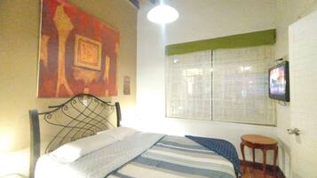 A bedroom with a bed, a television mounted on the wall, a window with blinds, and a painting on the wall.