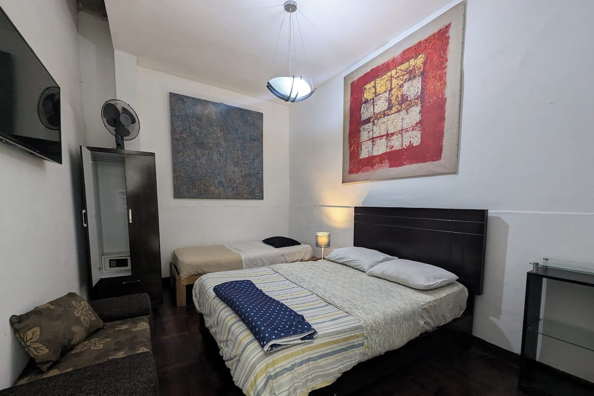 A bedroom with a bed, two bedside tables, a lamp, a fan, a large abstract painting, and a wardrobe.