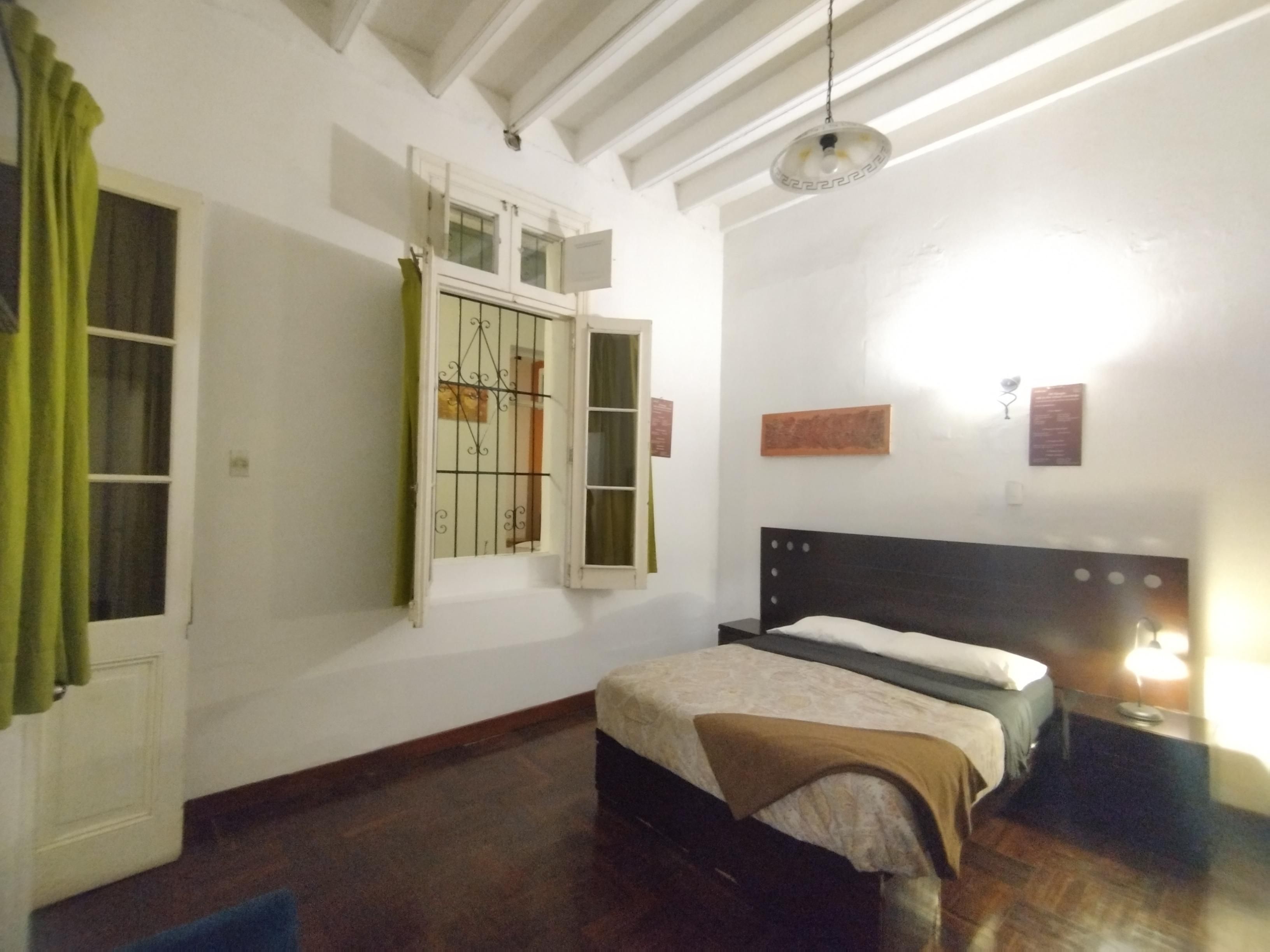 Une chambre à coucher avec un lit, une fenêtre avec des rideaux, un luminaire fixé au mur et un ventilateur de plafond.
