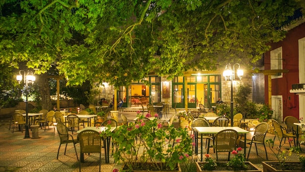 An outdoor cafe with tables and chairs, surrounded by greenery and illuminated by street lamps. - Oasis Exclusive  (Parga)
