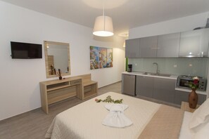 A modern hotel room with a bed, a wooden console table, a TV, a mirror, a painting, a kitchenette, and a vase with flowers. - Oasis Exclusive  (Parga)