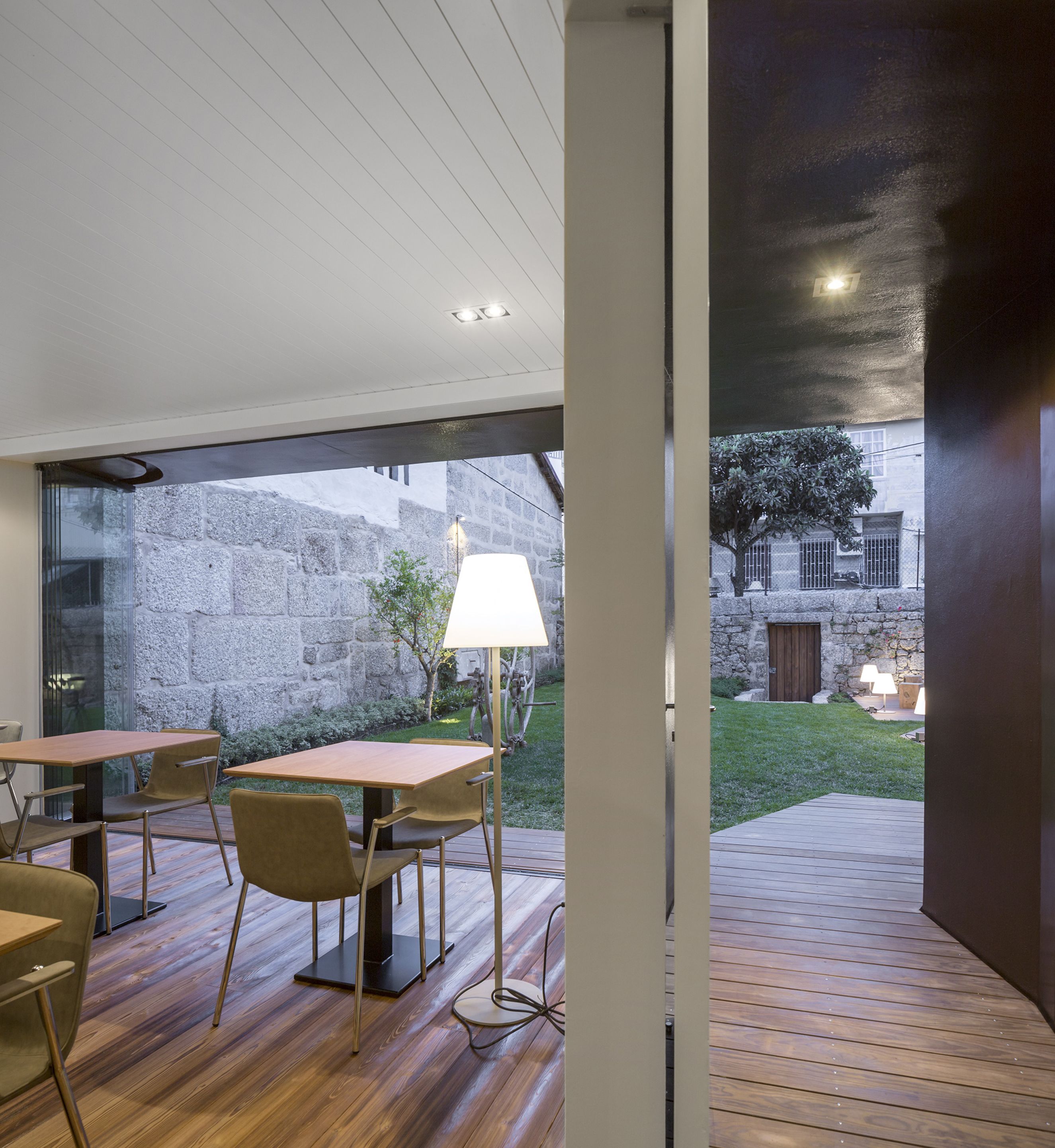 An interior space with a wooden floor, a dining area with tables and chairs, a lamp, and a view of a stone wall and greenery outside.