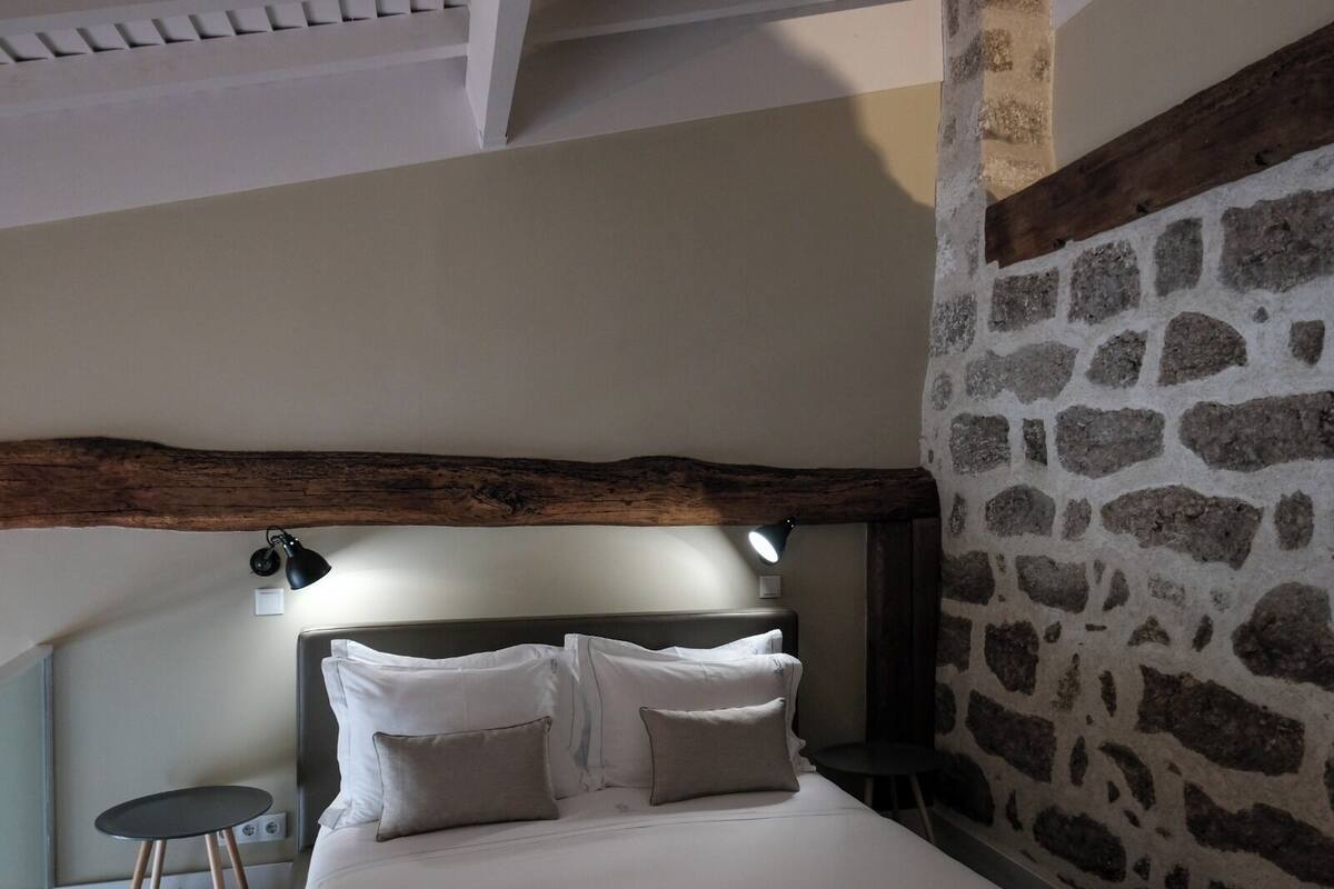 A bedroom with a stone wall, a wooden beam ceiling, a bed with white bedding, and two bedside lamps.