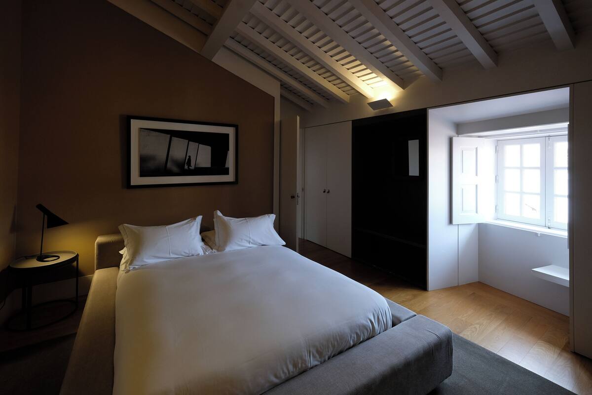 A bedroom with a large bed, a framed picture on the wall, a nightstand, and a window with blinds.
