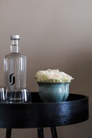 Una mesa con una botella transparente, un jarrón de cerámica con flores blancas y dos vasos.