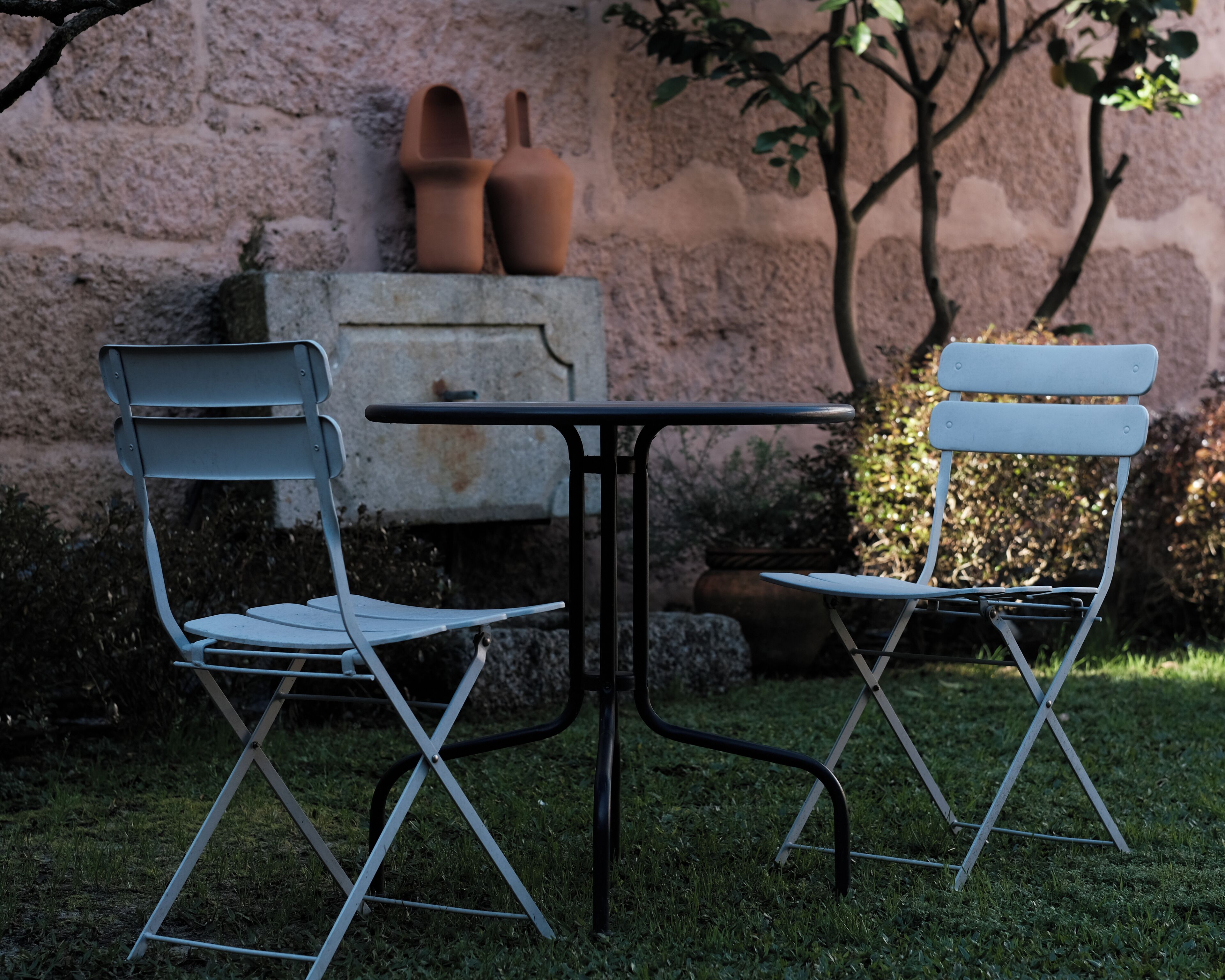 A small outdoor seating area with two metal chairs and a table, set against a stone wall with terracotta pots.