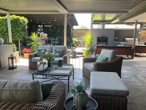 Een overdekt terras met rieten meubels, een bijzettafeltje en potplanten.