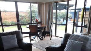 Un salon avec deux canapés en cuir noir, une table basse en bois sur laquelle est posé un vase bleu rempli de fleurs, et un coin repas à l’arrière-plan.