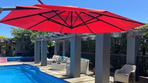 Un espace piscine agrémenté d’un parasol rouge, de meubles en osier et d’une structure en forme de pergola.
