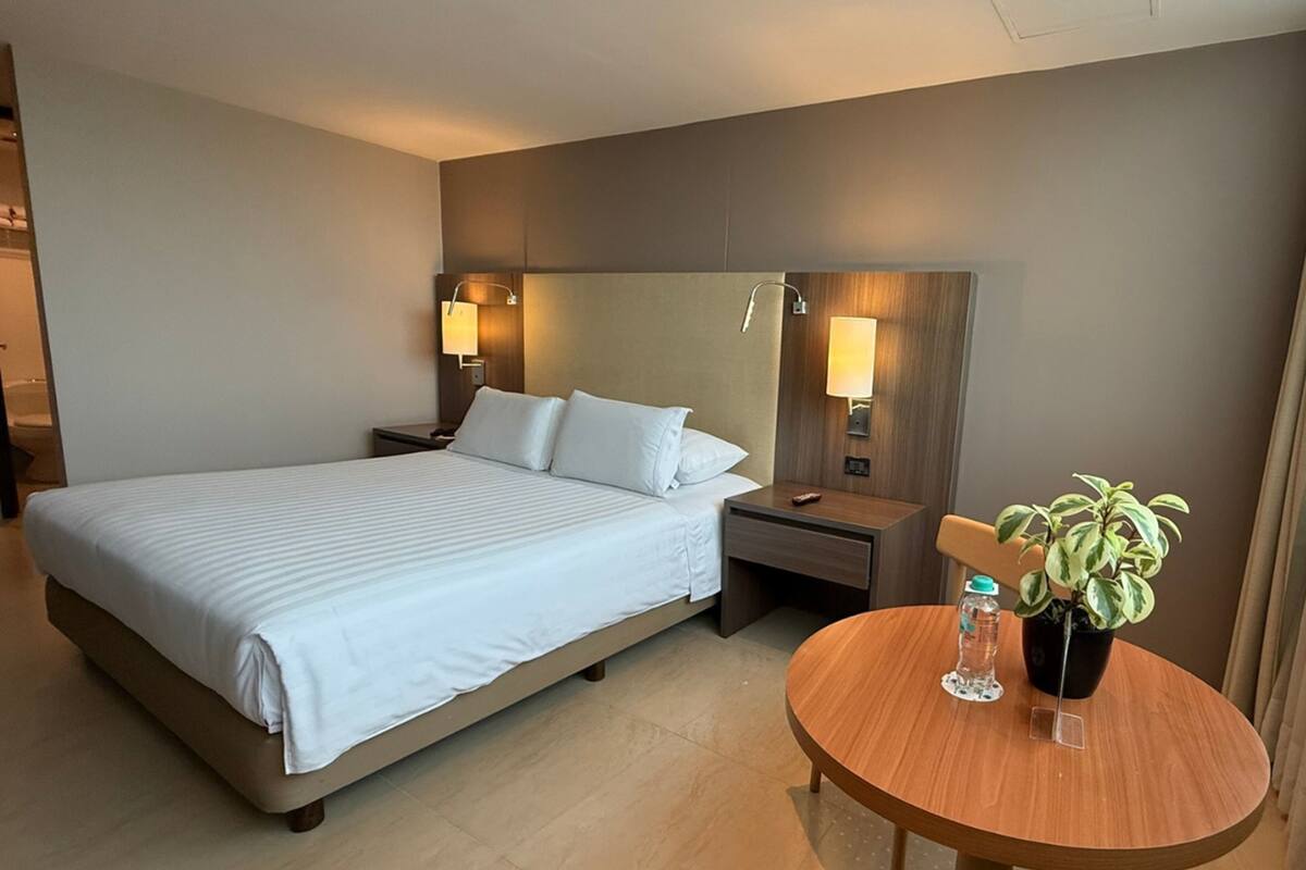 Ein Hotelzimmer mit Bett, Nachttischen, einem runden Holztisch und einer Zimmerpflanze sowie Wandleuchten.