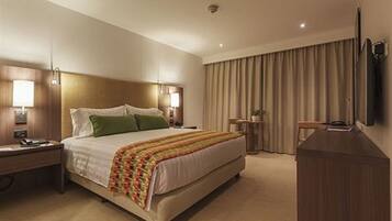 A hotel room with a large bed, bedside lamps, a desk, a flat-screen TV, and a large window with curtains.