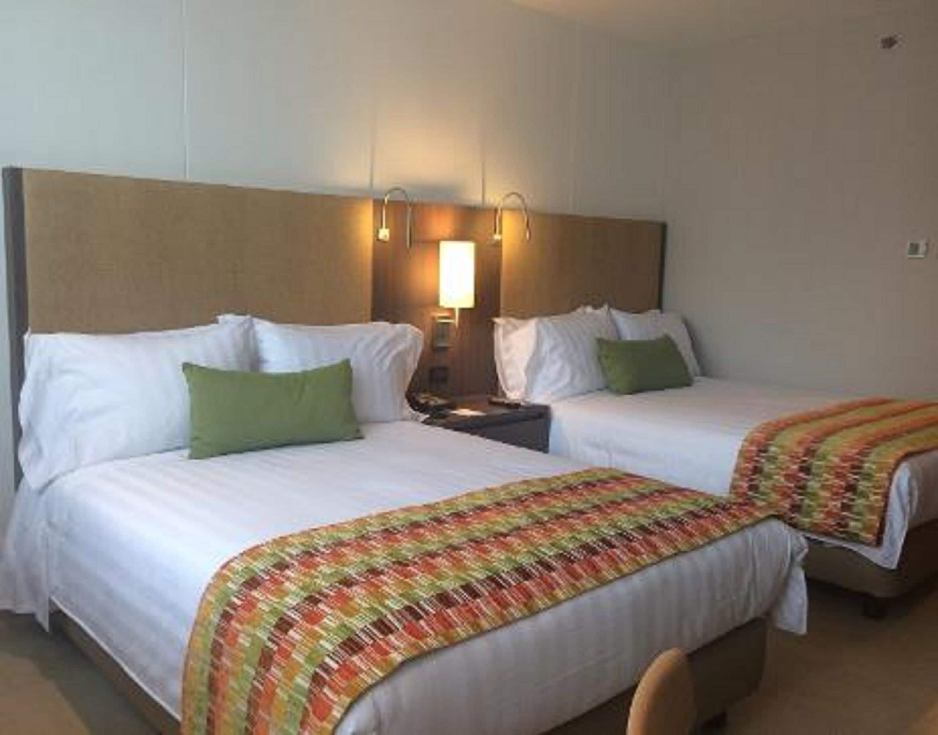 Habitación de hotel con dos camas, cada una con ropa blanca y almohadas verdes. Hay una mesita de noche con una lámpara entre las camas.