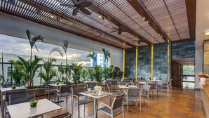 A spacious indoor dining area with multiple tables and chairs, a high ceiling with wooden beams, and large windows offering views of buildings and palm trees. - Estelar Villavicencio Hotel & Centro de Convenciones (Villavicencio)