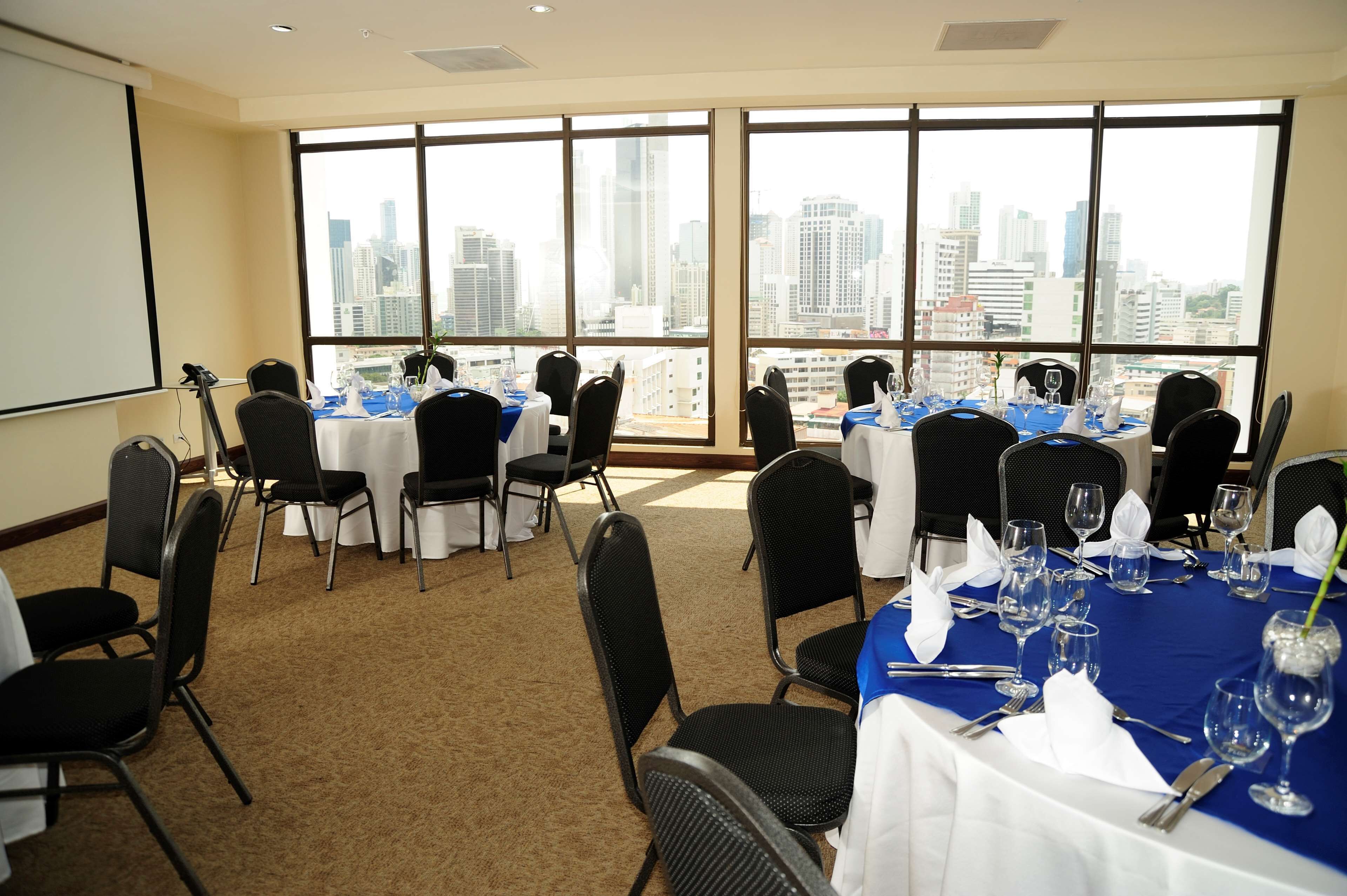 A conference room with multiple tables set for dining, large windows with a cityscape view, and a projector screen on the wall.