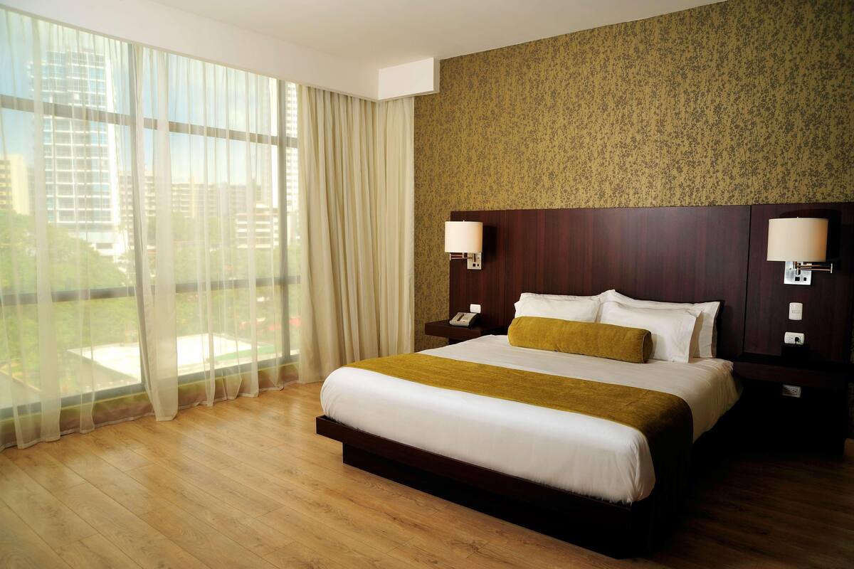 Une chambre d’hôtel avec un grand lit, deux tables de chevet avec des lampes, une vue sur la ville grâce à de grandes fenêtres et un mur décoré d’un motif.