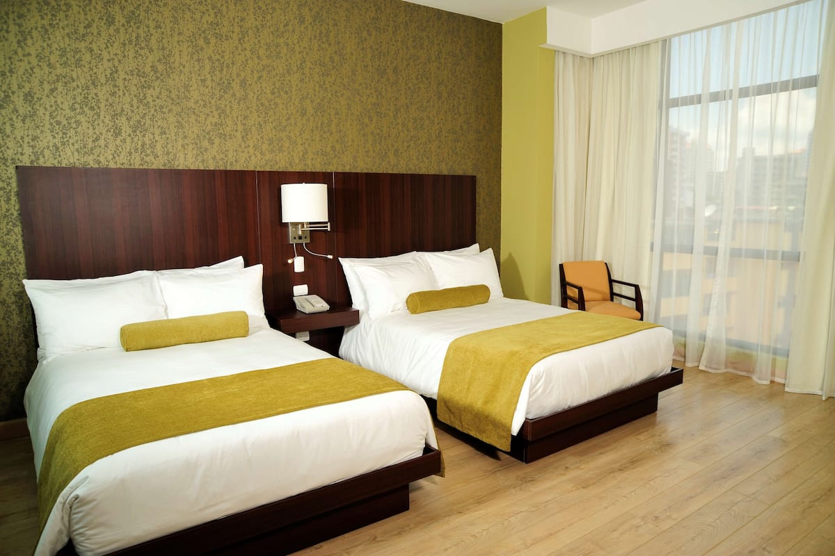 Une chambre d’hôtel avec deux lits, une tête de lit en bois, une table de chevet avec une lampe et une vue sur la ville à travers des rideaux transparents.