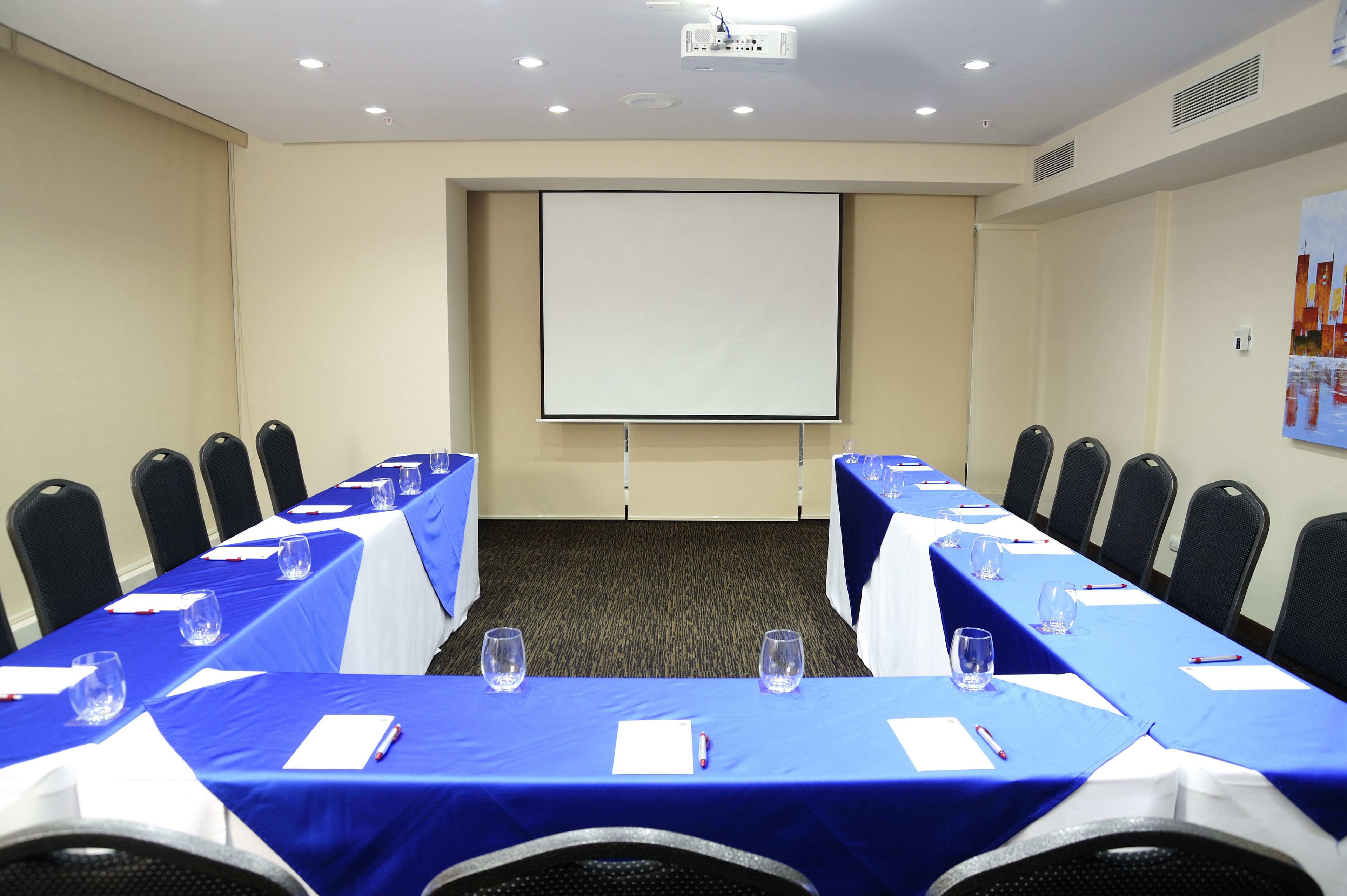 Un salón de conferencias con una larga mesa, sillas, una pantalla de proyector y suelo alfombrado.