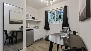 Cozinha compacta com armários brancos, micro-ondas e uma pequena mesa de jantar com cadeiras. Uma janela com cortinas pretas é visível, e um quadro emoldurado está pendurado na parede.