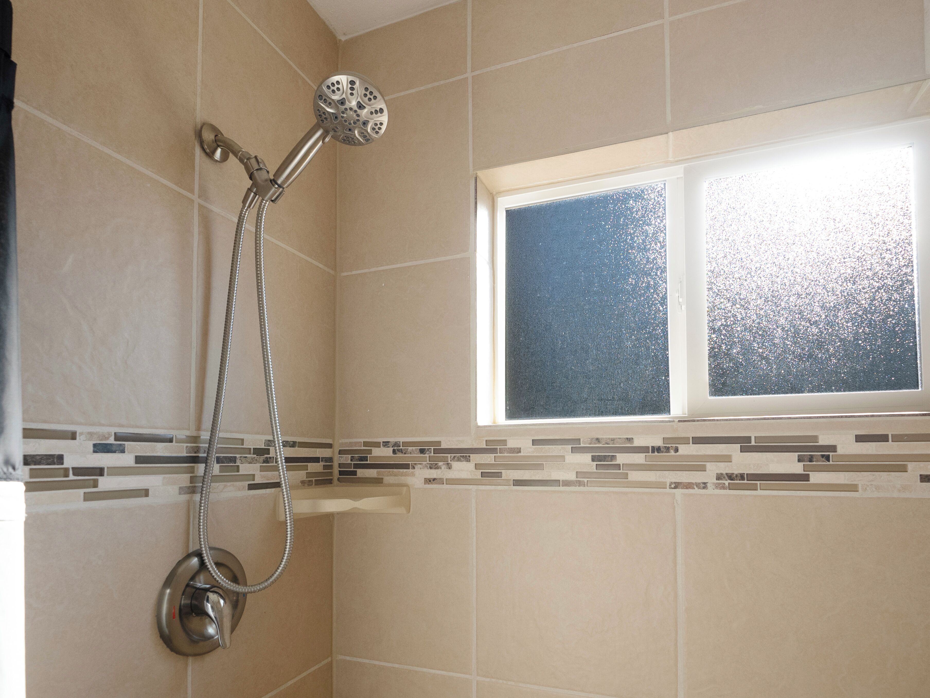 A shower with a handheld showerhead and a wall-mounted faucet, a window with a frosted glass, and a tiled wall with a decorative border.