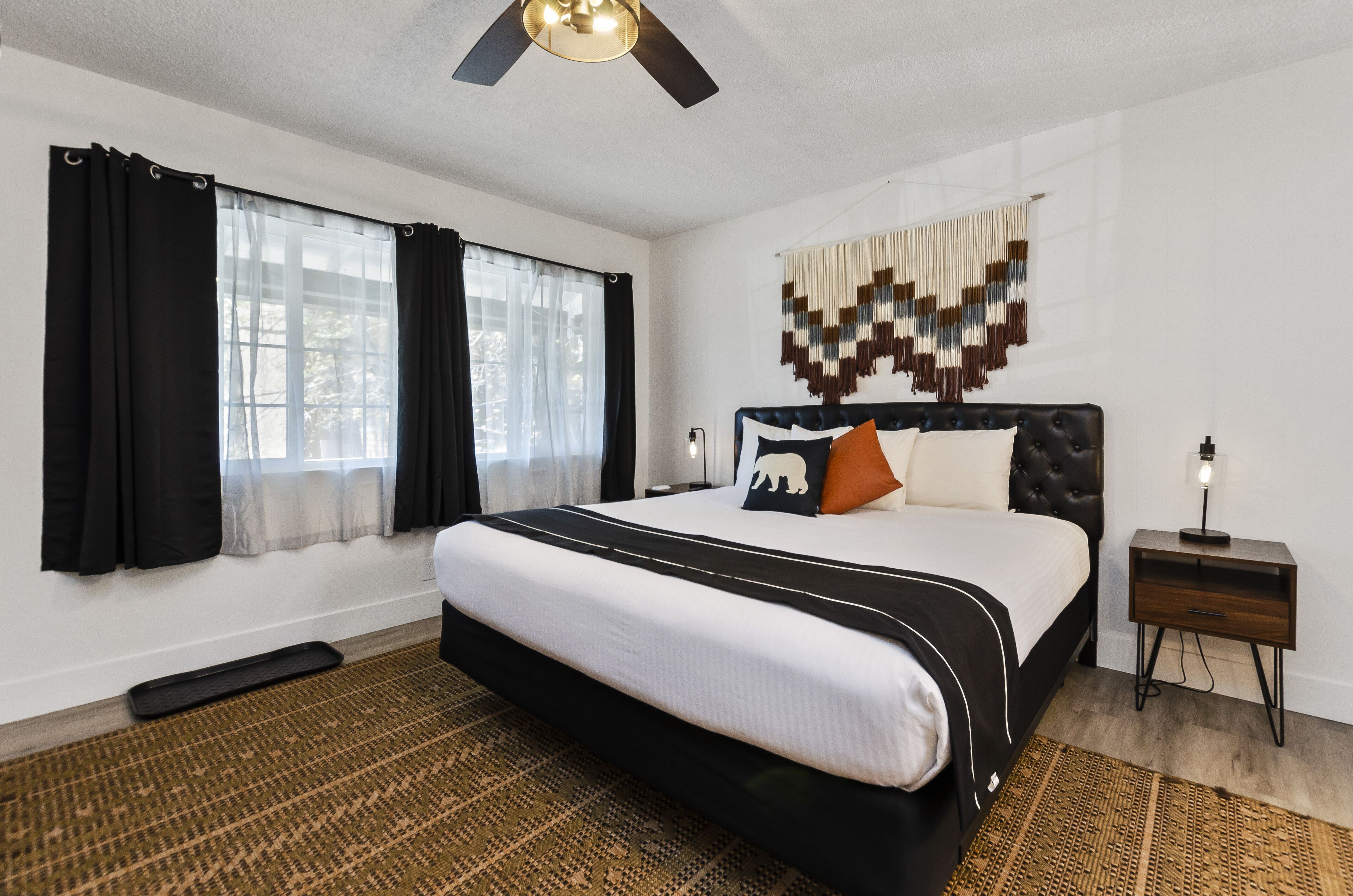 A bedroom with a large bed, a ceiling fan, a bedside table with a lamp, and a decorative wall hanging.